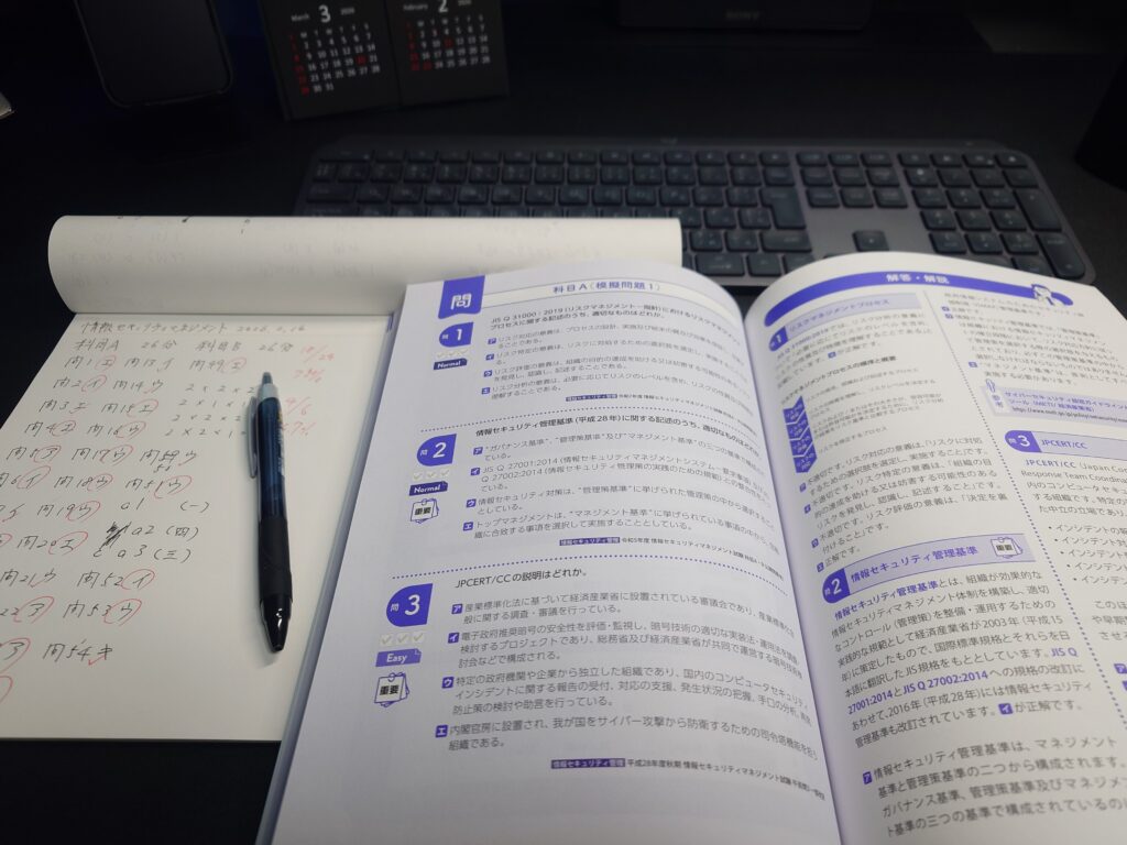 情報セキュリティマネジメント試験の問題集と手書きの解答ノート、ペンが机の上に並んでいる学習風景の写真